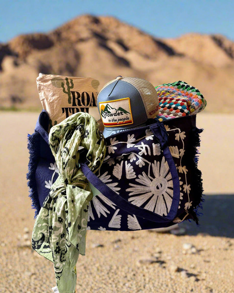 A blue tote bag with white floral patterns sits on desert ground. It contains a rolled blanket, a hat, a scarf, and a brown paper bag labeled “Road Trip.” Mountains are in the background.