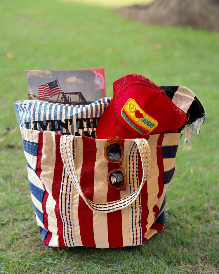 A red, white, and blue striped tote bag sits on grass, holding a rolled towel, a magazine, a red cap, black sunglasses, and a black-and-white striped cloth.