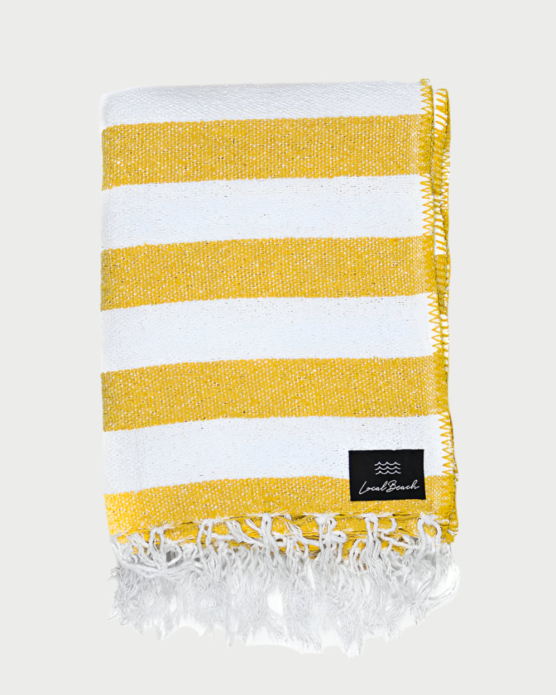 A folded XL LOCAL BEACH Baja Beach Blanket featuring wide yellow and white horizontal stripes, fringed tassels at one end, a crochet stitch border, and a small black “LOCAL BEACH” logo patch on the lower right corner.