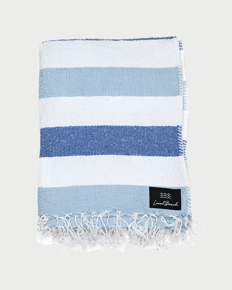 A folded, fringed LOCAL BEACH Baja Beach Blanket with wide blue and white stripes and a black woven label with white text in one corner, shown against a light background.