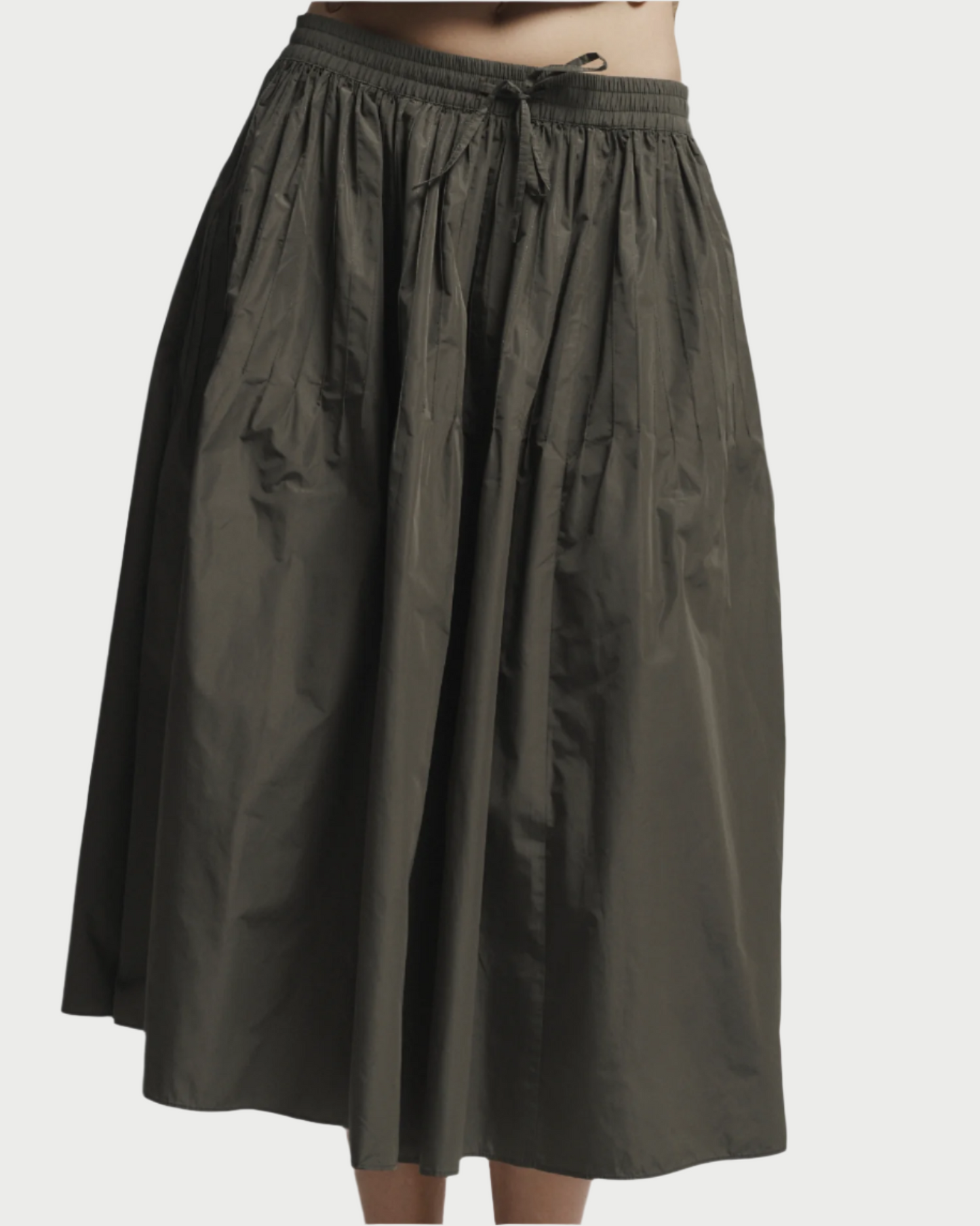 A person wears the Eva Midi Skirt by Saint Art NY in dark olive green, featuring a gathered waistband with waist ties. This high-waisted, ankle-length skirt has a loose fit and a slightly crinkled, flowy texture.