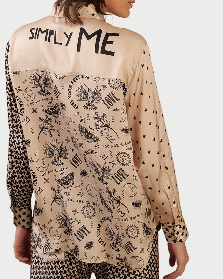 A woman wears the ME369 Scarlett Shirt, a beige button-down with black graphic hearts, eyes, stars, and positive words. The upper back features bold "SIMPLY ME" text for standout style.