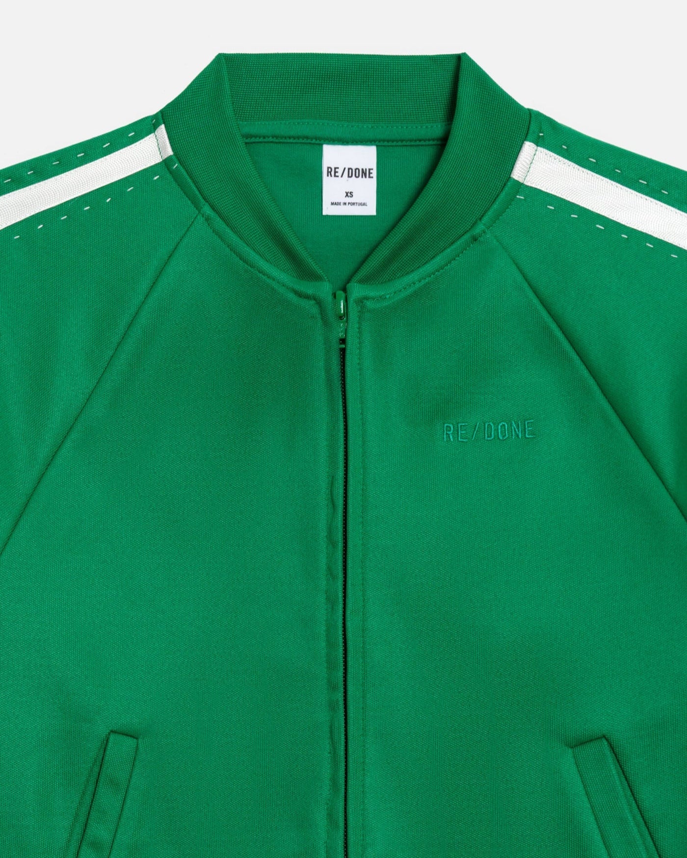 The Sunland Track Jacket by RE/DONE is green with white shoulder stripes featuring small stitched holes, a "RE/DONE" collar label, and the brand embroidered on the left chest for a sporty look.