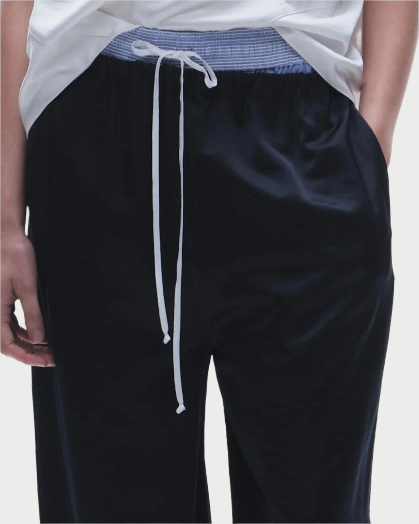 A person wears Saint Art NY’s Paige Pull-On Pant—loose-fitting black travel pants with a blue elastic waistband and white drawstring—paired with a white t-shirt. Only the lower torso and hands are shown.