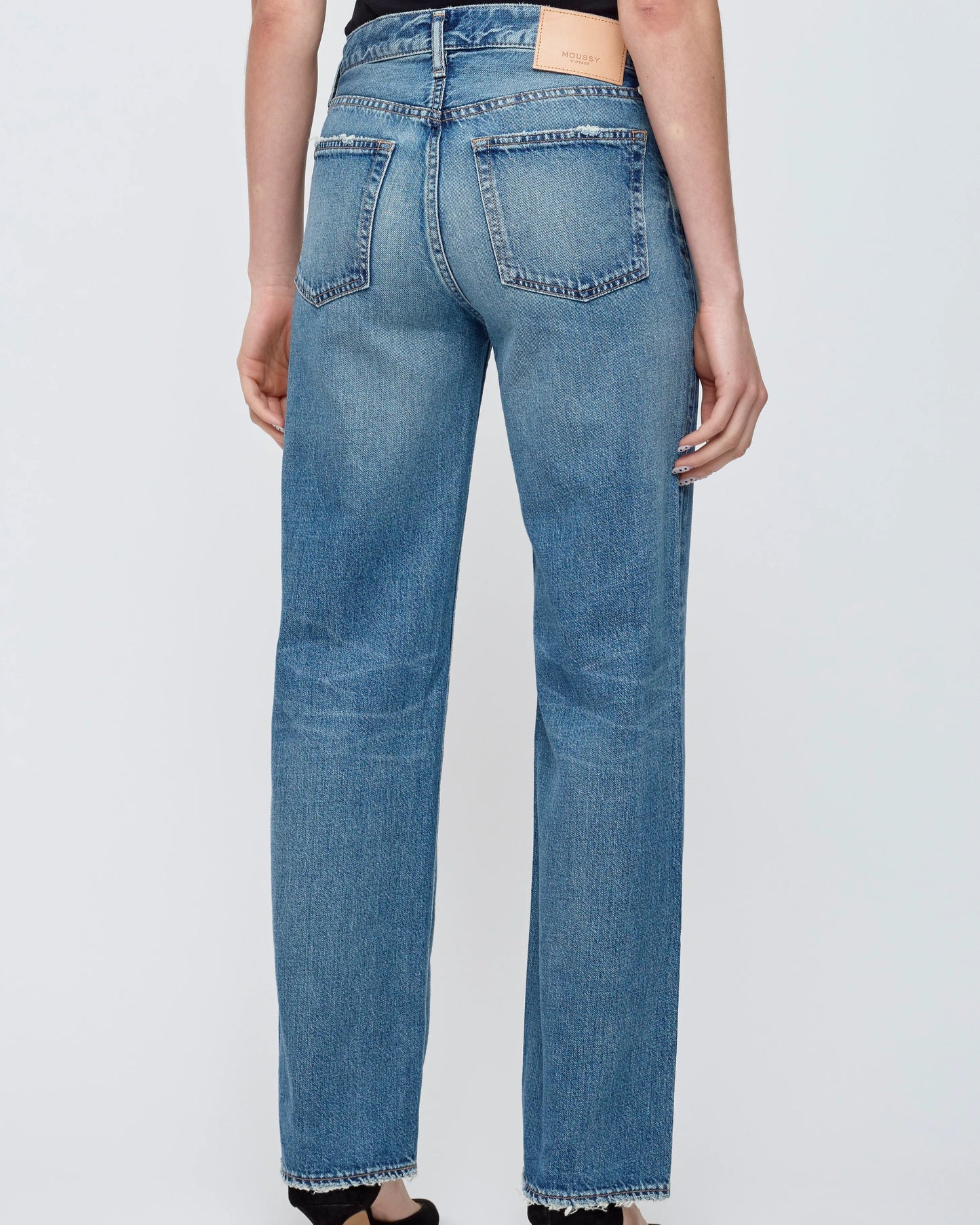 A person is seen from the back wearing Moussy’s Millsboro Straight Low Jeans in Japanese cotton denim, featuring straight legs, two back pockets, a leather waistband patch, and a raw hem against a plain background.