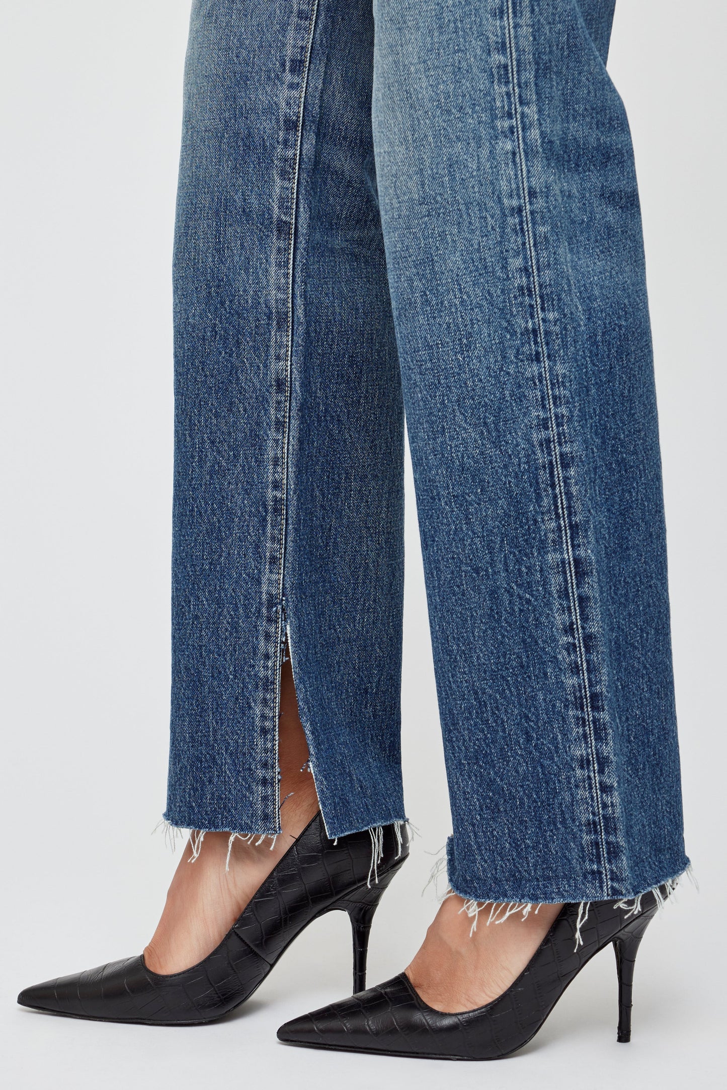 A person wearing Moussy Gleason Straight Leg Jeans with frayed hems and front slits, paired with black pointed-toe heels, stands against a plain white background.