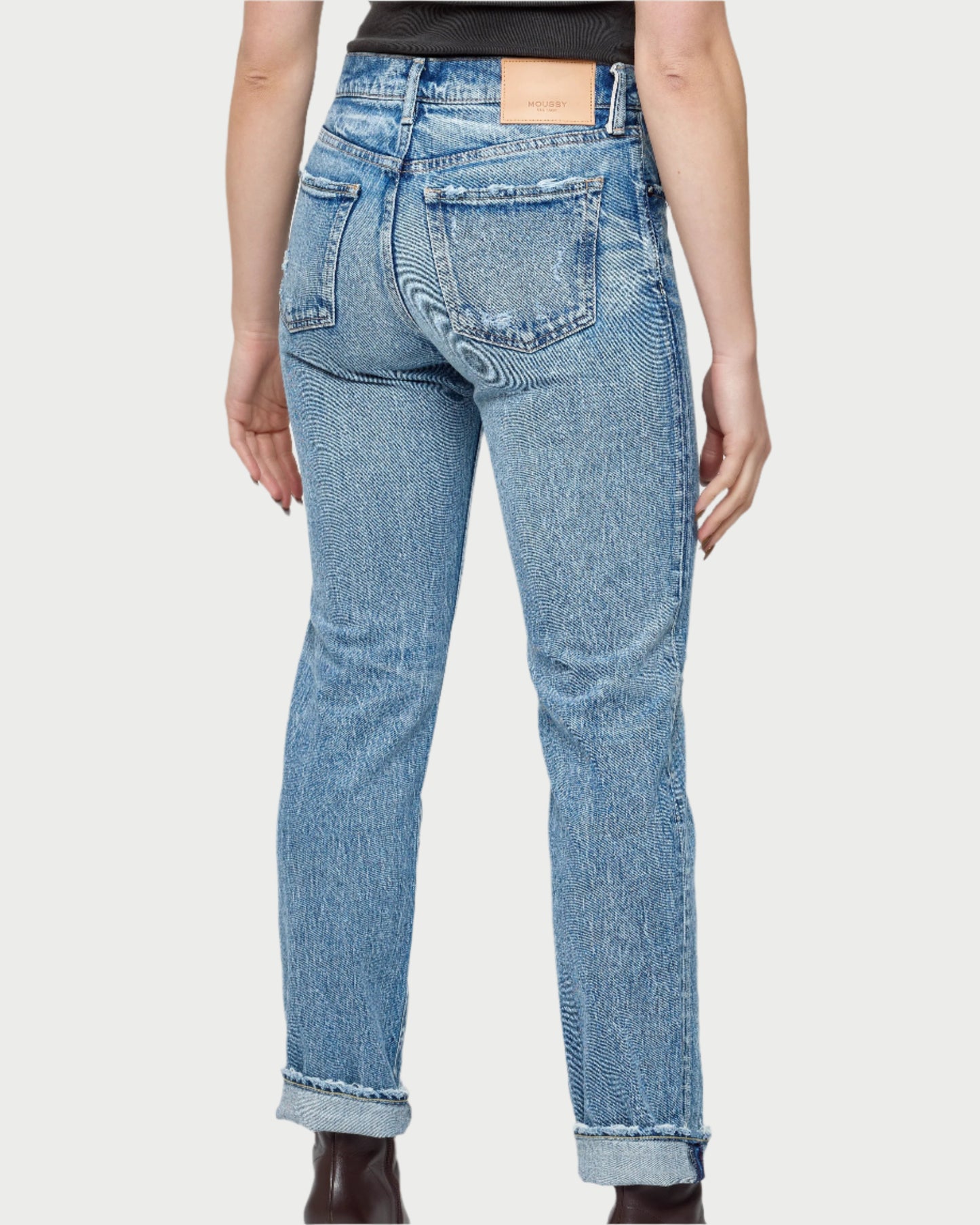 A person is seen from the back wearing Moussy Farmingdale Slim Straight Jeans in light blue Japanese denim with rolled cuffs, a tan leather patch on the waistband, styled with a black top and brown ankle boots.