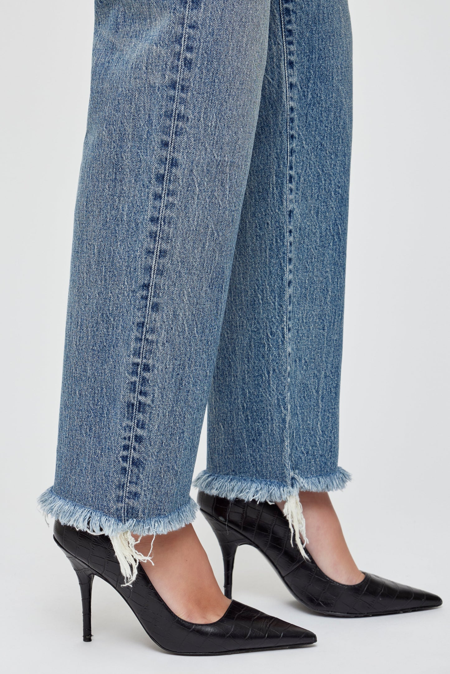 A person wears Moussy Massapequa Straight Leg Jeans with frayed hems and black pointed-toe heels, standing against a light background.