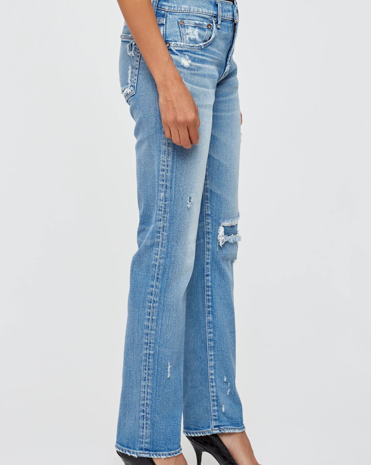 A person stands against a plain light background wearing light blue MOUSSY Fairview Slim Straight Jeans with rips on the thigh and knee, paired with black high-heeled shoes.