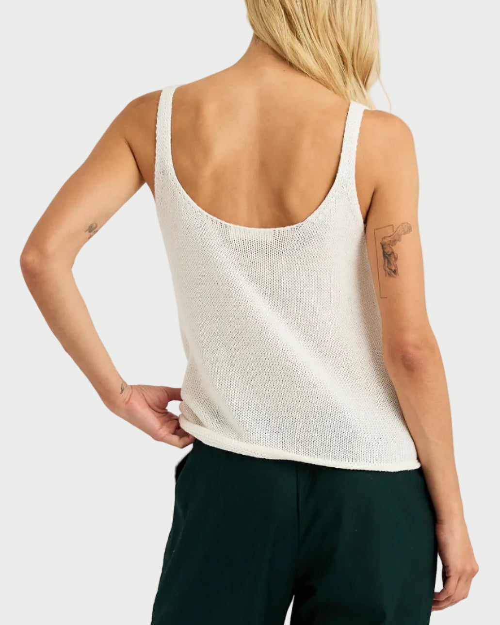 Person wearing a white sleeveless top and dark green pants on a light gray background