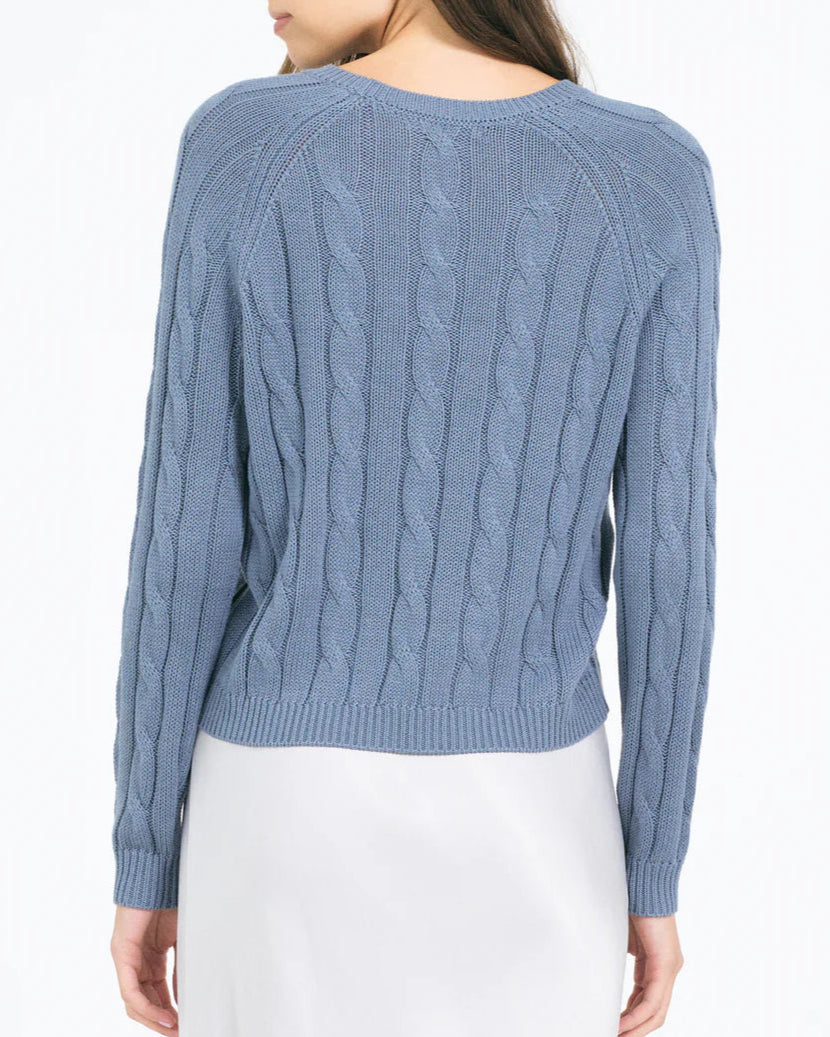 A woman is seen from the back in a light blue One Grey Day Raleigh Cable Pullover, featuring soft cotton-viscose fabric, cable knit details, and a ribbed hem, paired with a white skirt against a plain white background.