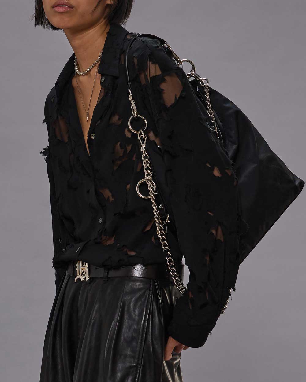Person wearing a Slim Abstract Print Shirt by R13, black leather pants, silver chain belt, layered necklaces, and carrying a large black bag with silver chain straps over their shoulder against a plain gray background.