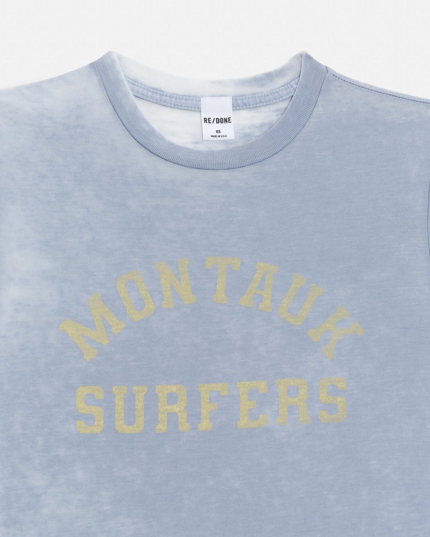 The RE/DONE Montauk Tee is a light blue vintage-style shirt with a faded burnout finish and “MONTAUK SURFERS” in yellow arched text on the chest. The inner tag features the RE/DONE branding.