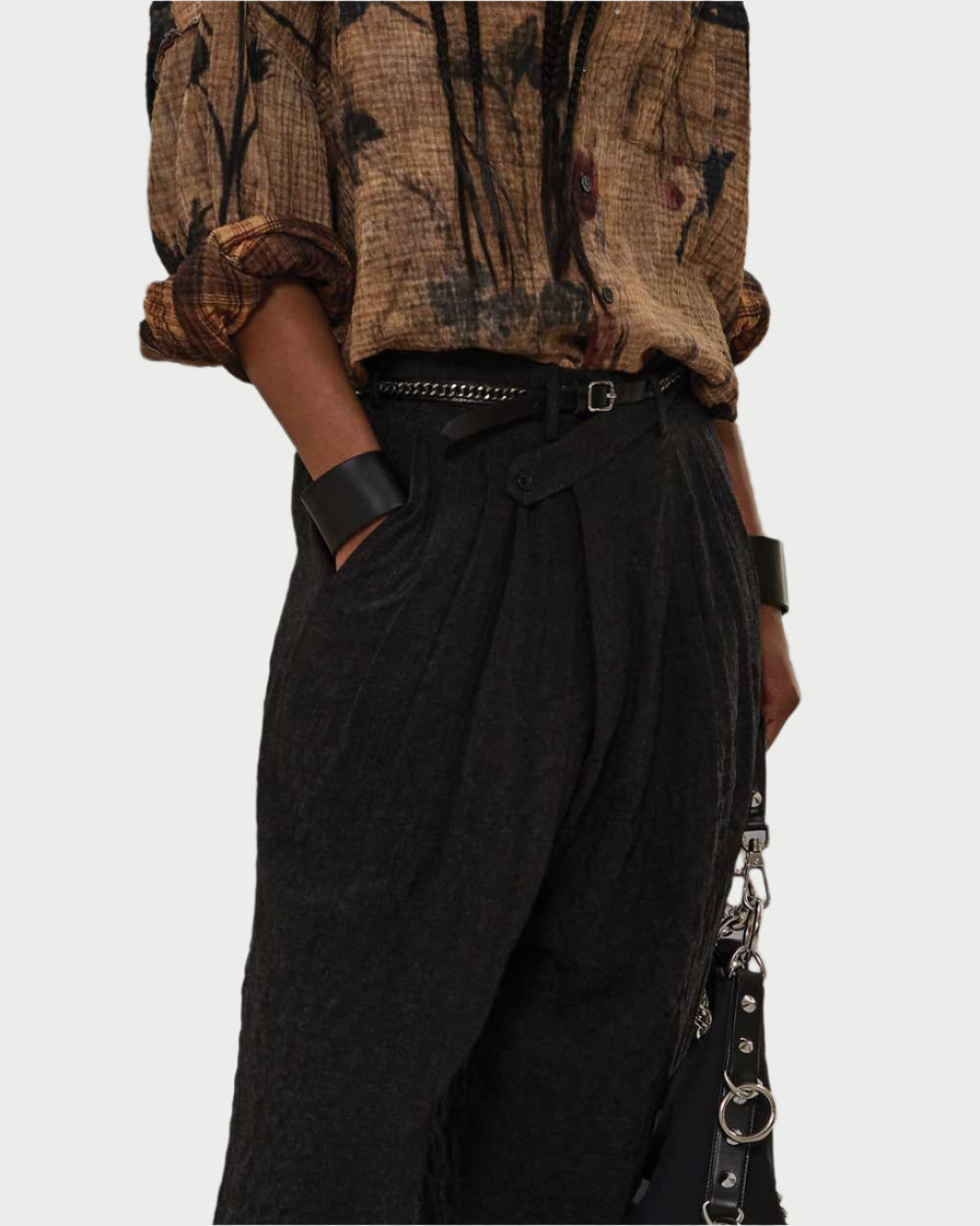 Someone wearing a crinkled brown floral shirt tucked into R13 Triple Pleat Crossover Trousers with a belt, styled with a chunky black bracelet and a black handbag featuring metal details.