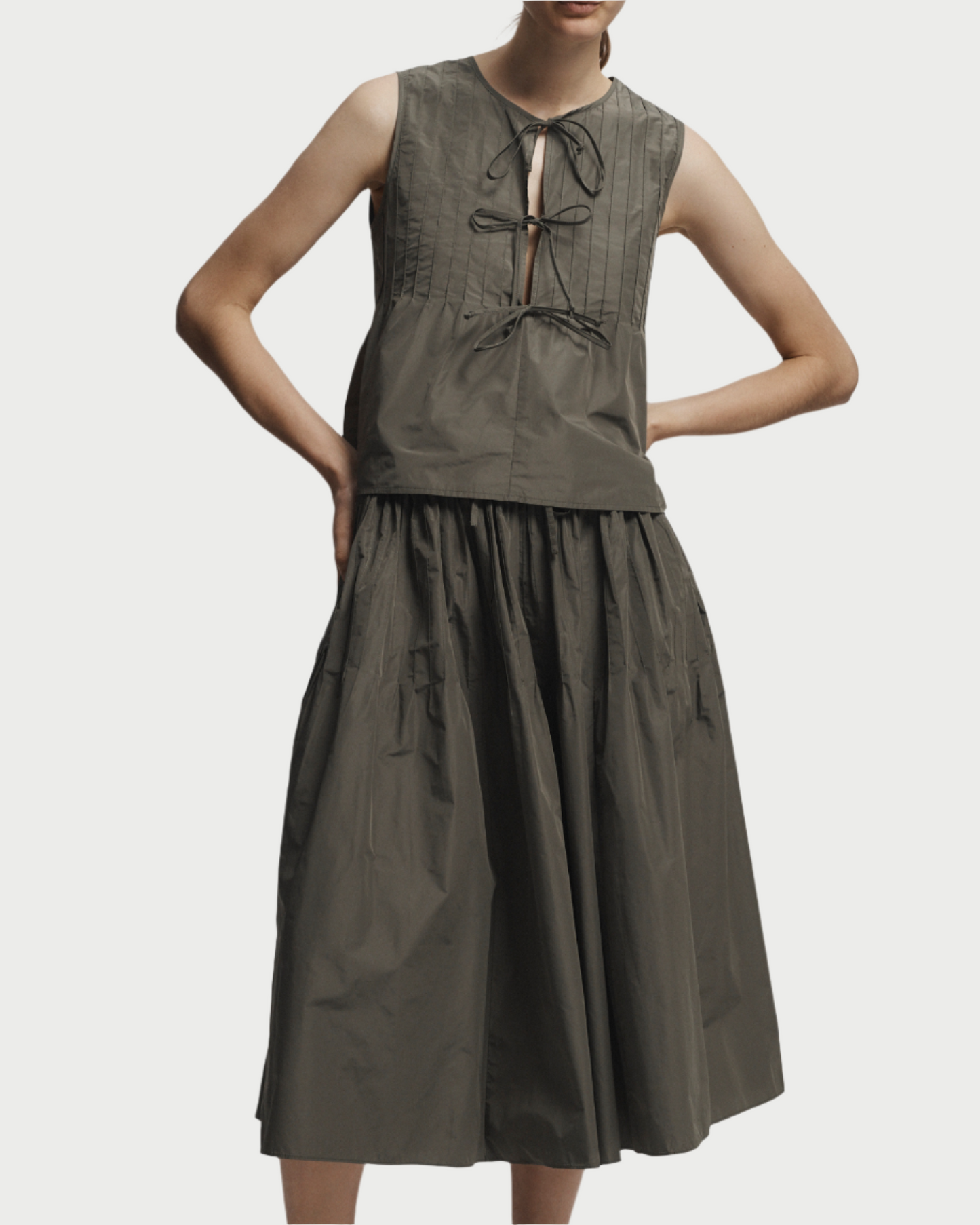Wearing the Saint Art NY Nina Tie Front Top, a sleeveless pleated olive top with front ties, and a matching full midi skirt, they add a playful touch while posing with one hand on their hip and black strappy sandals.