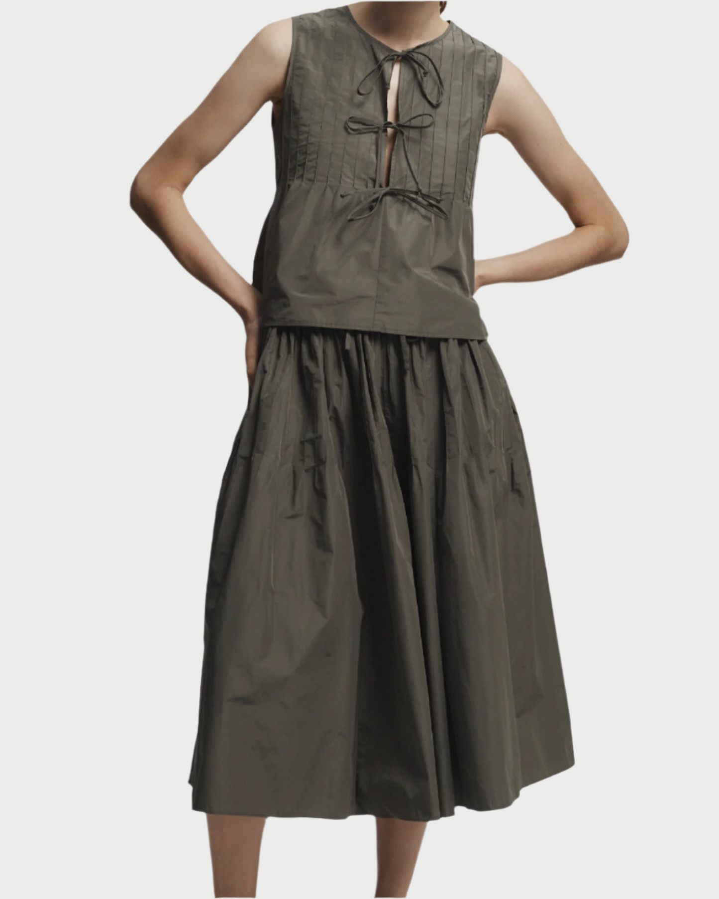 A person wears the Saint Art NY Eva Midi Skirt in dark olive green, styled sleeveless with waist ties and a pleated bodice. Their arms are bent and face is not visible.