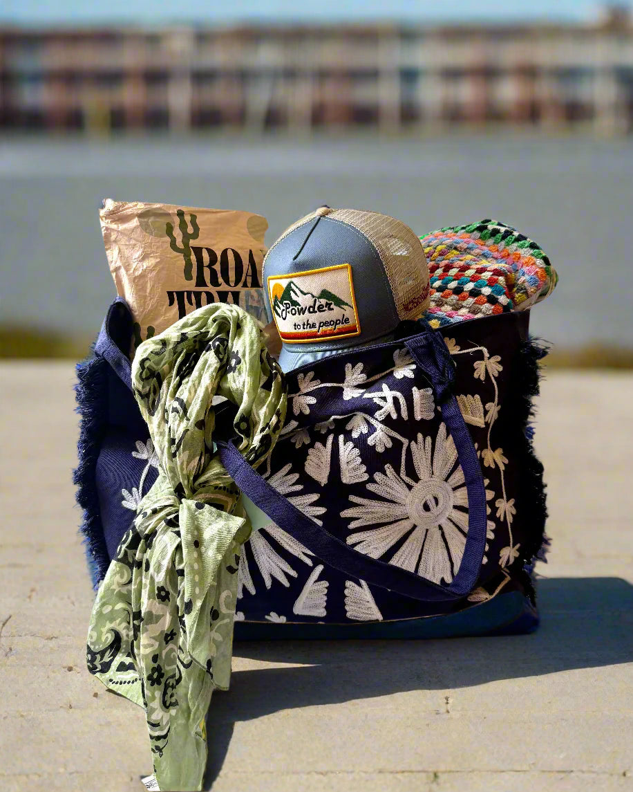 A DEBBIE KATZ Boho Bag in navy blue with white floral patterns rests on a ledge, holding a tan blanket, colorful crochet, green scarf, and gray trucker hat with a "Powder to the people" patch.