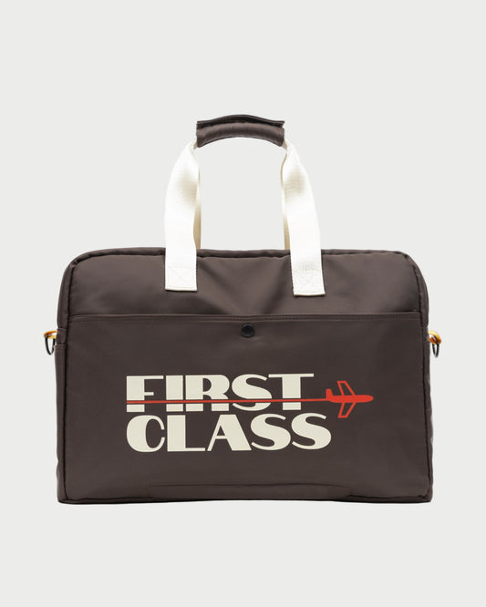 The Oncept Olympia Travel Bag by Oncept is a dark brown carry-all with white handles, displaying "FIRST CLASS" in bold white letters and a red airplane graphic integrated into the text on the front.