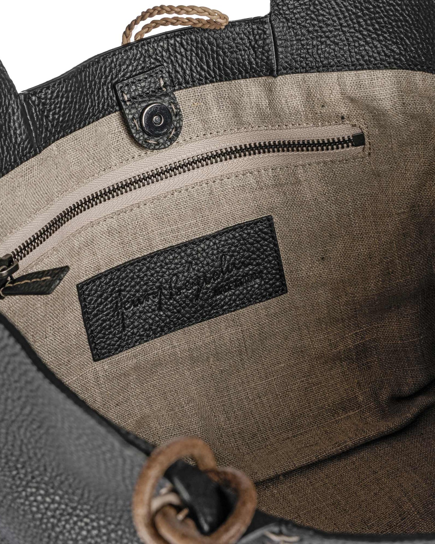 The Medium Muflone Tote by Henry Beguelin is a calfskin leather handbag with beige fabric lining, a zipper pocket, and black label. Handmade in Italy, it features a button closure and braided handle, showcasing timeless elegance and quality craftsmanship.