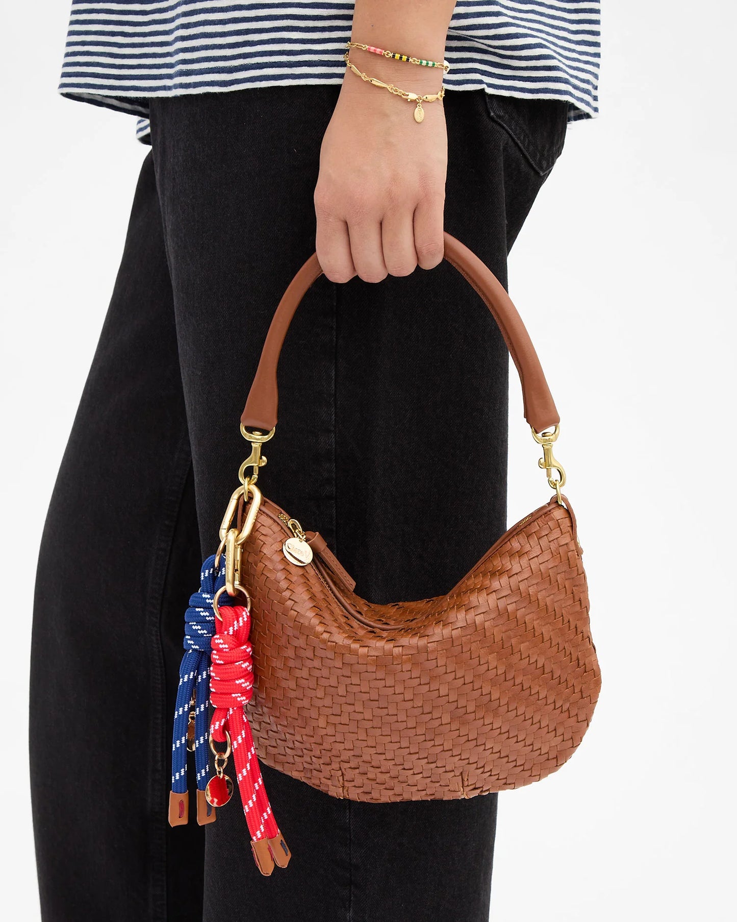 A person wearing a striped shirt and black pants holds a small, brown woven handbag with a curved handle, adorned with red and blue keychains—including the Clare V. Barrel Knot Fob by CLARE V.—as an accessory. The person also wears a gold bracelet with small charms.