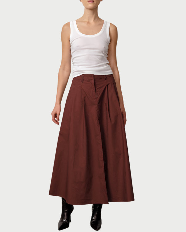 Woman wearing a white tank top and brown long skirt against a plain background