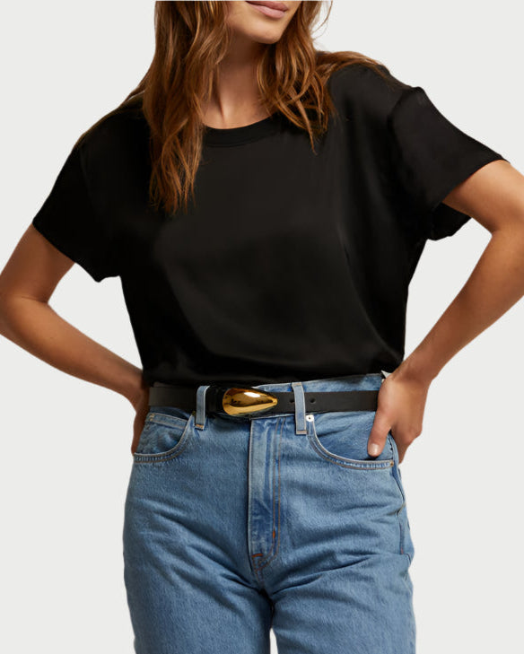 A person wearing a black Delphine Satin Crewneck Tee from perfect white tee and blue jeans, with hands on hips. A stylish belt featuring a gold buckle adds an elevated touch against the plain background.