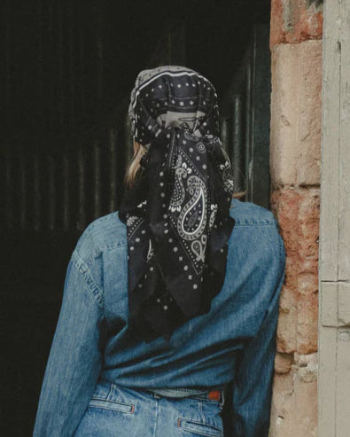 A person in a blue denim jacket and jeans, near a weathered brick wall, wears the Maison Lecomte Flament Rodeo Scarf—a cotton voile scarf with a black bandana pattern—covering their head and facing away from the camera.