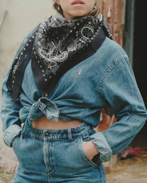 A person wearing a knotted denim shirt, high-waisted jeans, and the Maison Lecomte Flament Rodeo Scarf with a bandana pattern stands with one hand in their pocket. Their face is partially out of frame.