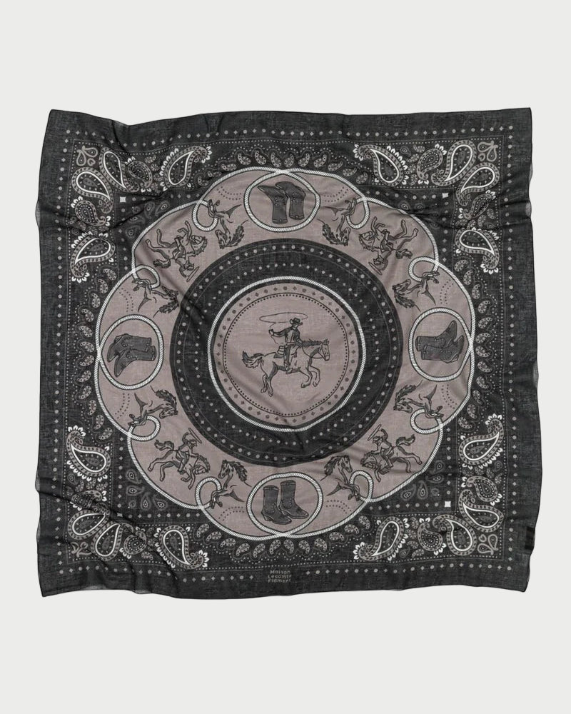 The Maison Lecomte Flament Rodeo Scarf features a black and gray bandana pattern with a central cowboy on horseback, horses, boots, hats, and paisley borders. Its relaxed look comes from the slightly wrinkled fabric.