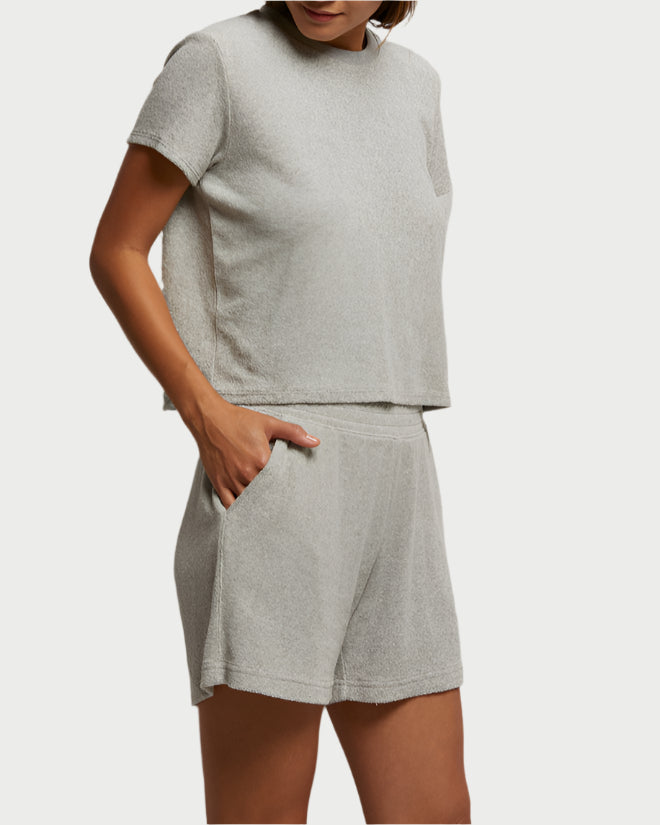 A person in a light gray, short-sleeve loop terry top and Emily Loop Terry Shorts by perfectwhitetee stands with one hand in a pocket. The casual and comfortable outfit has a relaxed fit, accentuated against the plain background.