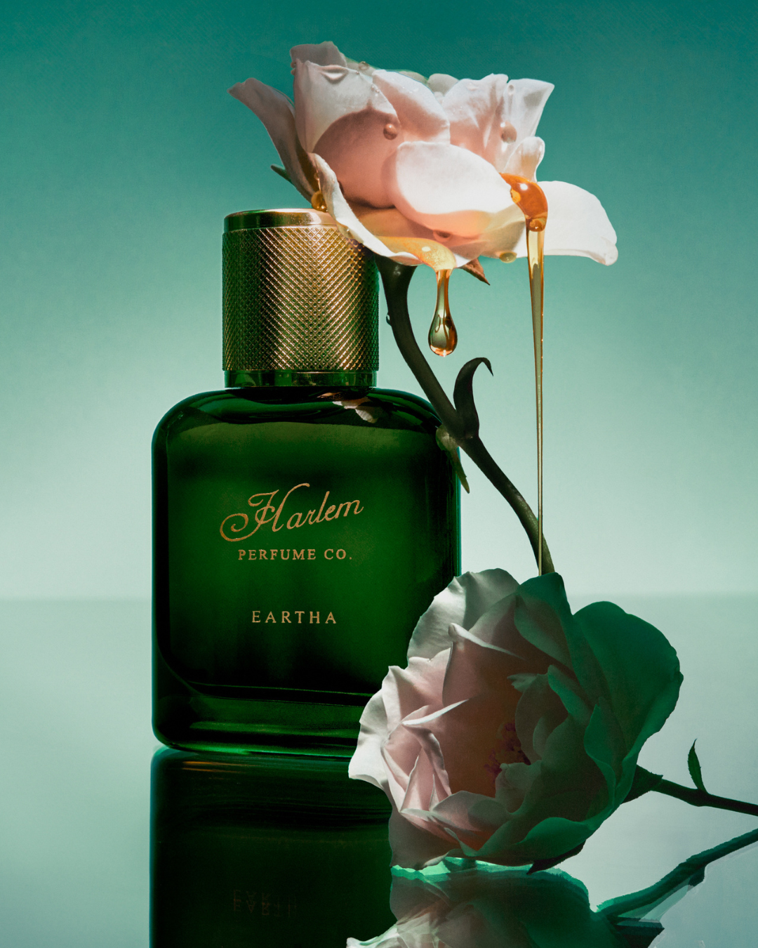 A green Harlem Perfume Co. EARTHA EAU DE PARFUM bottle stands by pale pink roses, one dripping golden liquid onto it—capturing the fragrance’s fearless sophistication against a teal background.