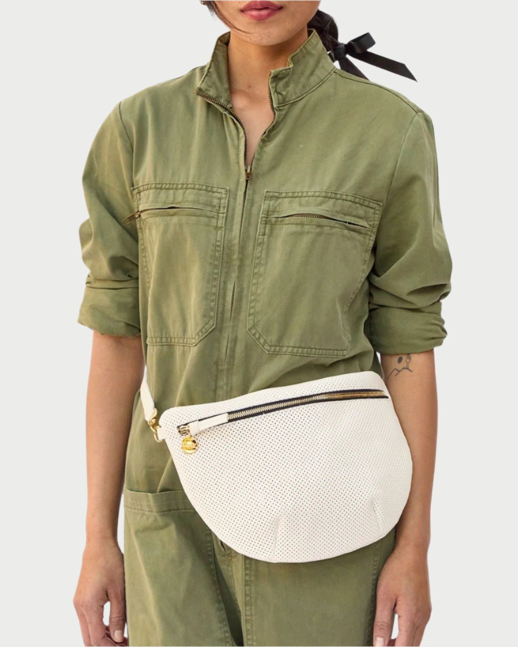 The person wears a green jumpsuit with rolled-up sleeves, showcasing the CLARE V. Grande Fanny Bag, featuring a black zipper and gold accents, elegantly positioned at the front. A black ribbon ties their hair, adding a touch of flair.