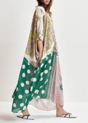 A woman stands against a plain background wearing the Essentiel Antwerp Hansas Printed Caftan, a flowy, oversized maxi dress featuring green and white polka dots, floral and geometric patterns, and a deep V-neckline, paired with flat white sandals.