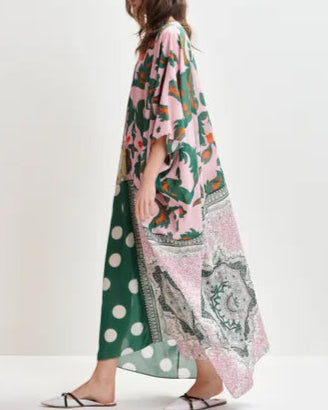 A person wears the Hansas Printed Caftan by Essentiel Antwerp—an oversized, colorful piece with mixed prints—paired with white slide sandals, standing against a plain background.