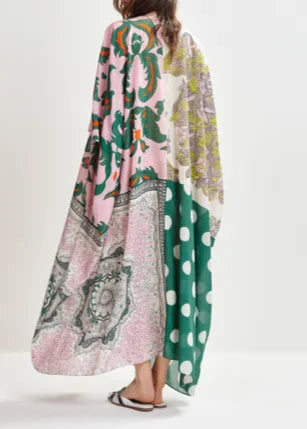 A woman stands facing away, wearing the Essentiel Antwerp Hansas Printed Caftan—a colorful, oversized dress with a deep V-neckline and mixed patterns in pink, green, white, and yellow. She completes the look with sandals.