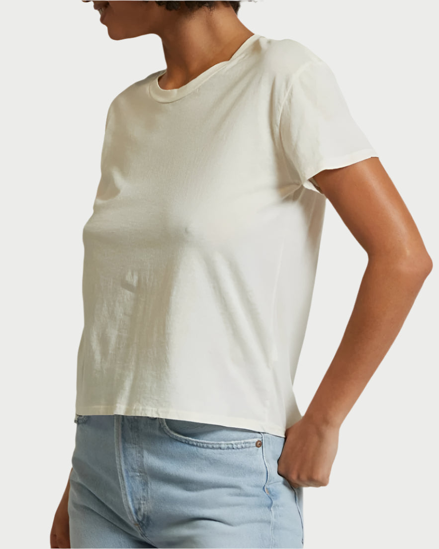 A person wearing a Bright Ivory Harley T-Shirt by perfectwhitetee and mid-rise denim stands with one hand in their pocket against a neutral background.