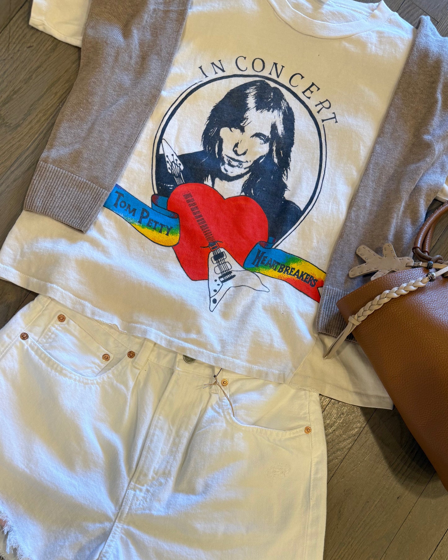 A flat lay features a Madeworn Tom Petty Tee in vintage fit with an illustration and colorful text, paired with a beige cardigan, 100% cotton white denim shorts, and a brown leather handbag with a decorative tag, all elegantly arranged on a wooden floor.