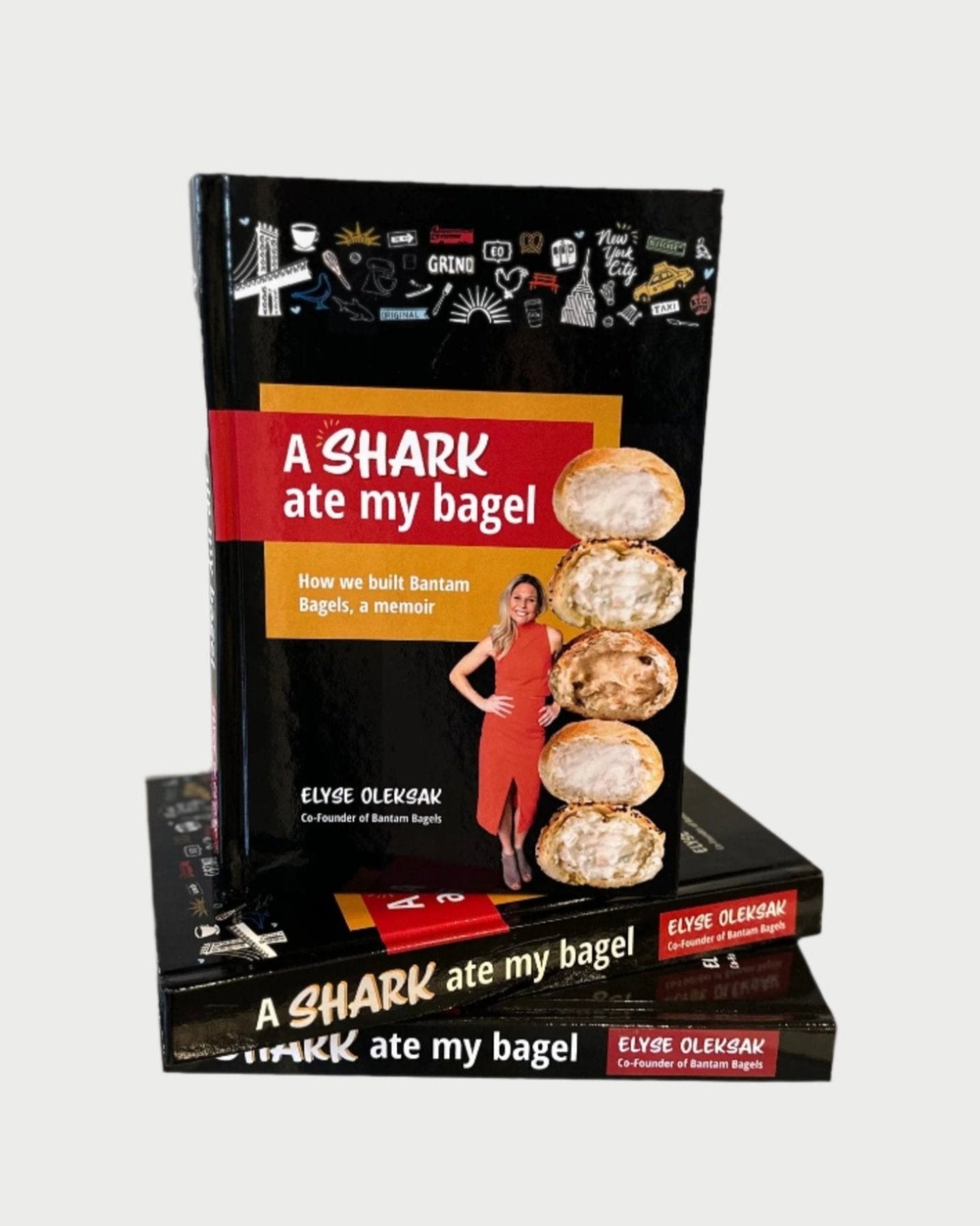 The book "A Shark Ate My Bagel" by Elyse Oleksak, co-founder of Bantam Bagels and Shark Tank alumna, features a cover with her beside large bagels. Two more copies lay flat beneath it.