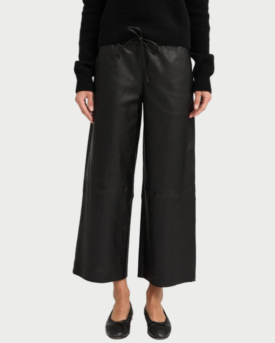 A person wearing SPRWMN Cropped Leather Baggy Pants, a black long-sleeve top, and black flat shoes stands against a plain light background.