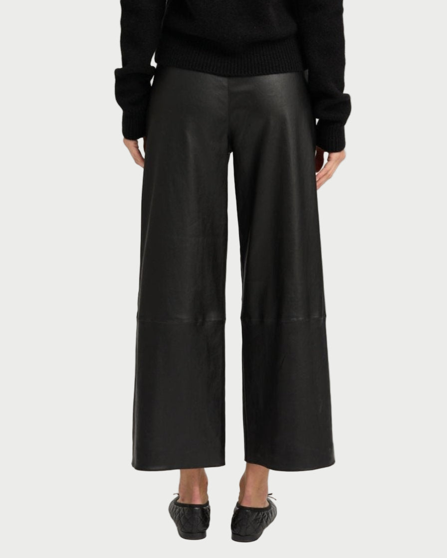 Someone wearing SPRWMN Cropped Leather Baggy Pants, a black sweater, and black shoes stands with their back to the camera against a plain light background.