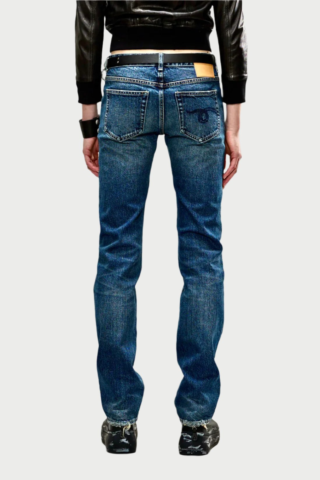 A person stands facing away, showcasing the back view of their outfit. They are wearing a black leather jacket, 100% cotton low-rise slim leg jeans with pockets, and black sneakers. The jeans have a light brown label on the waistband emblazoned with "Tobi Boy Jean," and the background is plain gray.