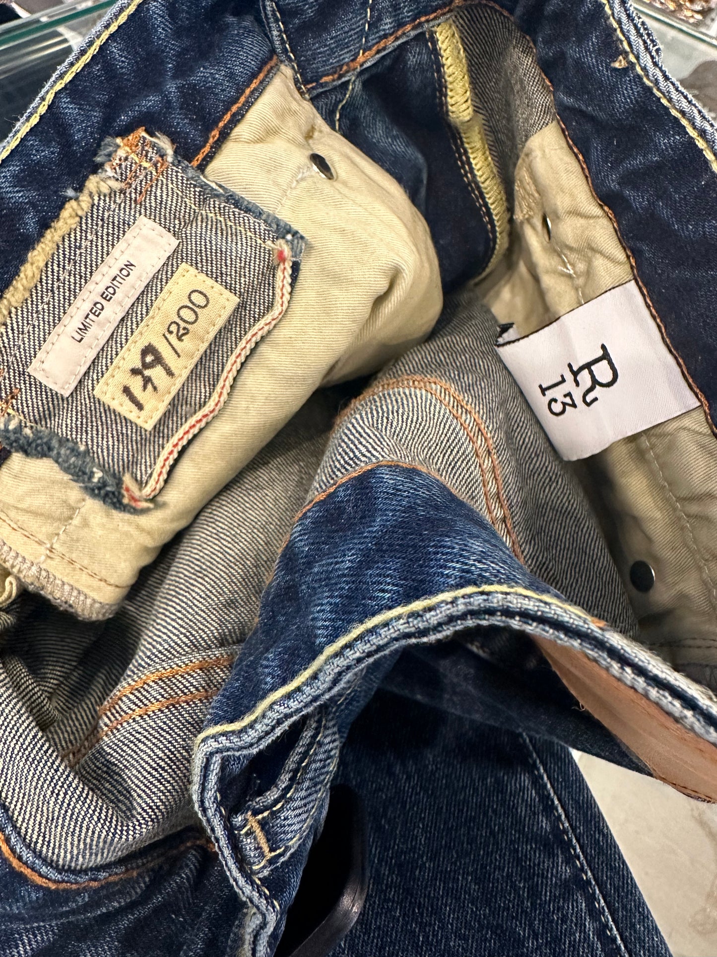 A close-up of the inside of a pair of blue denim jeans reveals a limited edition label, "17/200." Another white label displays the brand "R13." The jeans, identified as Tobi Boy Jean, feature tan-colored stitching and visible pocket linings.