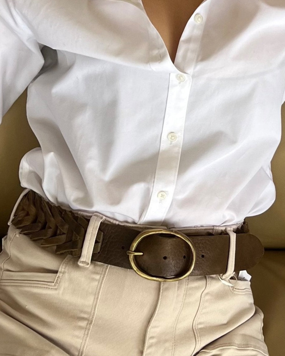 Wearing a white button-up tucked into beige pants, a person completes the casual, classic look with Motif56's Spike Leather Belt, featuring a versatile wide brown leather design, a round gold buckle, and subtle triangle link accents.