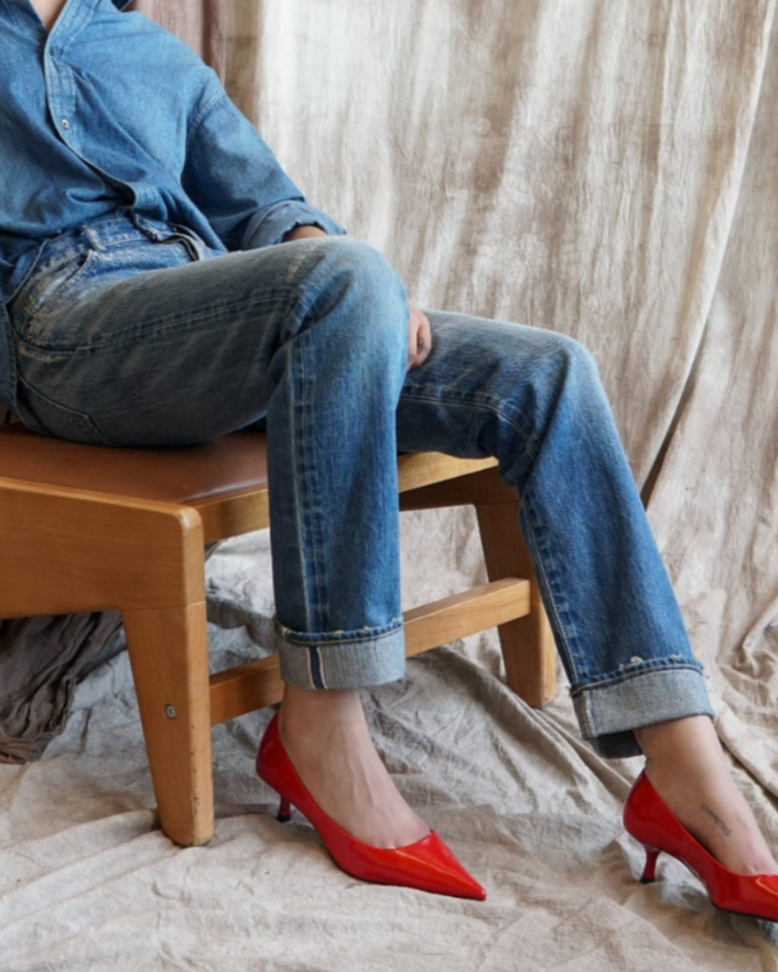 Seated on a wooden chair with their face hidden, a person wears Moussy’s Frederica Straight Leg Jeans in blue denim, paired with a matching blue denim shirt and bright red heels. The backdrop features draped light brown fabric.