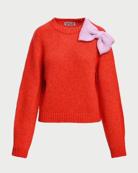 A bright orange-red ribbed knit sweater with a large light purple bow attached to the left shoulder, shown against a plain light gray background.