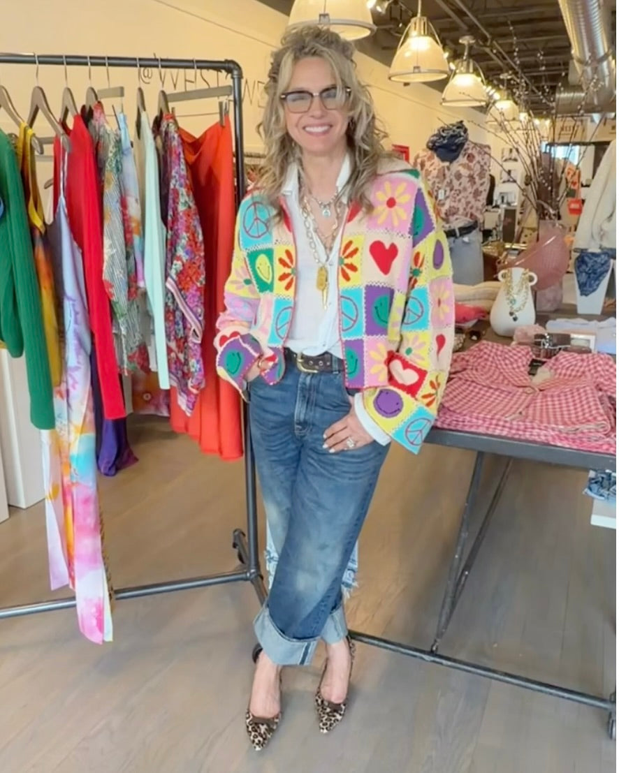 A smiling woman wearing glasses and the ME369 Miami Crochet Cardigan stands in a boutique surrounded by vibrant ME369 clothes and accessories.
