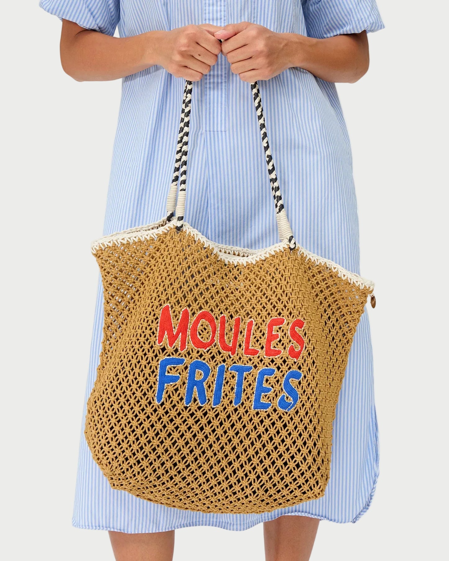 A person in a light blue striped dress holds the Clare V. Moules Frites L'Été Tote by CLARE V., which features "MOULES FRITES" in red and blue lettering and black-and-white rope handles.