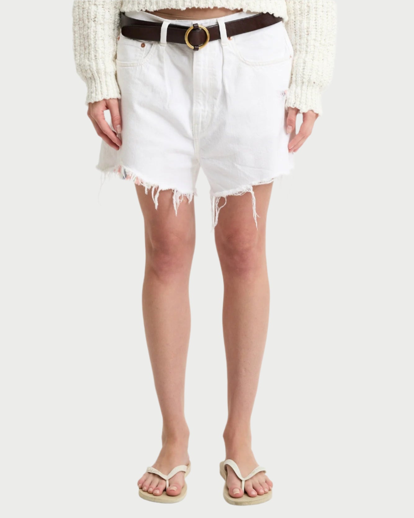 A person is wearing Denimist's Brooke Shorts in white with frayed edges, paired with a dark brown belt and a cream knit sweater. They complete the contemporary look with premium cotton white flip-flops, standing against a plain background.