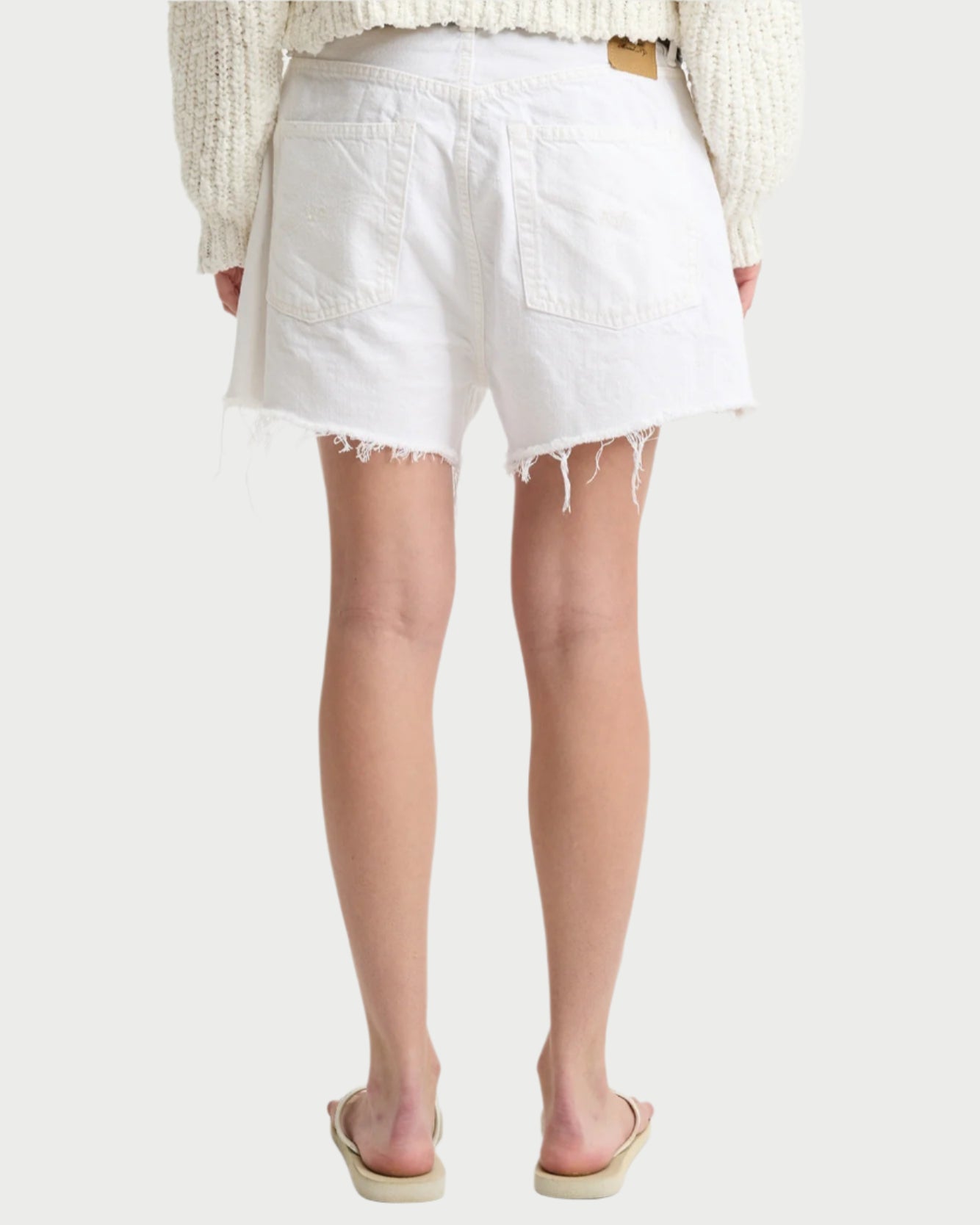 A person is seen from the back wearing Denimist's Brooke Shorts in white denim with frayed hems, paired with a cream knit sweater and beige flip-flops, all against a light background. Crafted from premium cotton, these shorts evoke contemporary style.