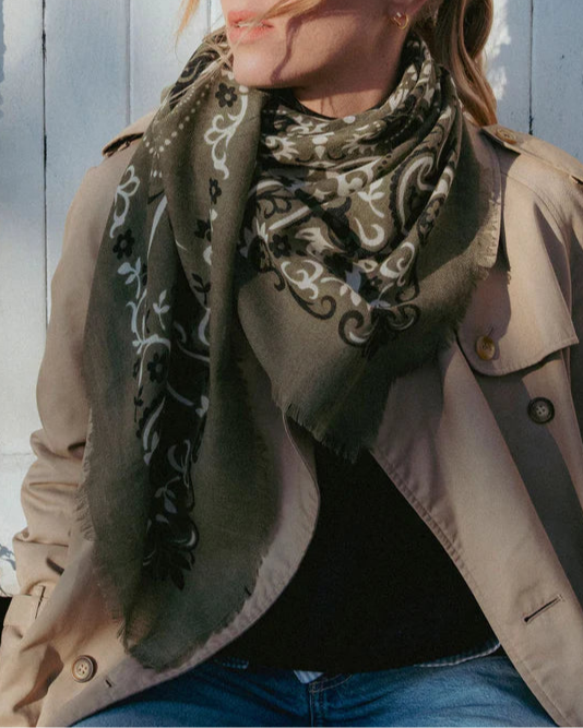 A person in a beige trench coat, black top, and blue jeans sits against a white wooden background, accessorized with a Maison Lecomte Flament Bandana Scarf Khaki.